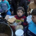 Palestinian children hold plates and containers while receiving food aid from charitable workers in Deir el-Balah, Gaza Strip during Ramadan. A young boy in a red and yellow shirt looks directly at the camera while others gather around food distribution pots.