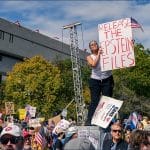 Woman standing above crowd holding large "Release the Epstein Files" sign at outdoor rally with protesters and additional signs visible in background