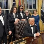 Donald Trump in tuxedo seated at Resolute Desk with Kennedy Center honorees including KISS members standing behind him in Oval Office