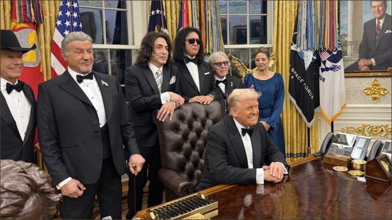 Donald Trump in tuxedo seated at Resolute Desk with Kennedy Center honorees including KISS members standing behind him in Oval Office