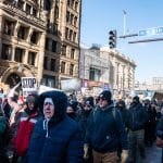Protesters in winter coats march past Masonic Temple in Minneapolis carrying signs reading We Will Not Comply With Tyranny and Stop ICE Terror