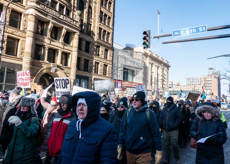 Protesters in winter coats march past Masonic Temple in Minneapolis carrying signs reading We Will Not Comply With Tyranny and Stop ICE Terror