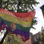 Rainbow pride flag with National Park Service arrowhead logo reading Stonewall National Monument Established 2016 flying over Greenwich Village New York City