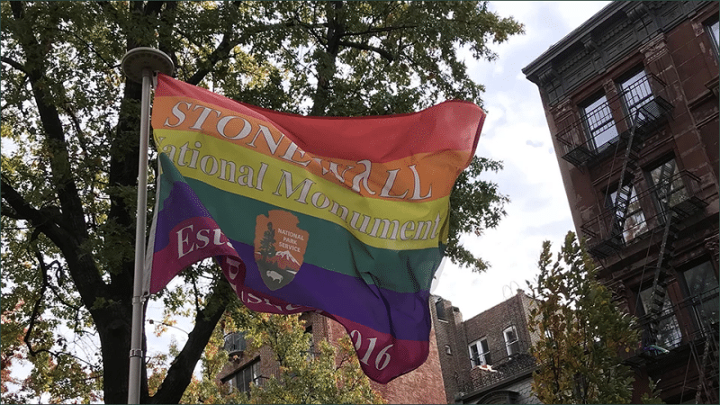 Rainbow pride flag with National Park Service arrowhead logo reading Stonewall National Monument Established 2016 flying over Greenwich Village New York City