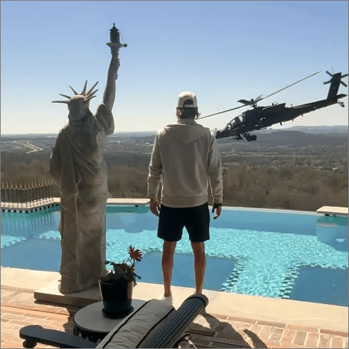 Kid Rock standing poolside at his Nashville estate watching an Army Apache helicopter hover near his swimming pool with a Statue of Liberty replica in the foreground