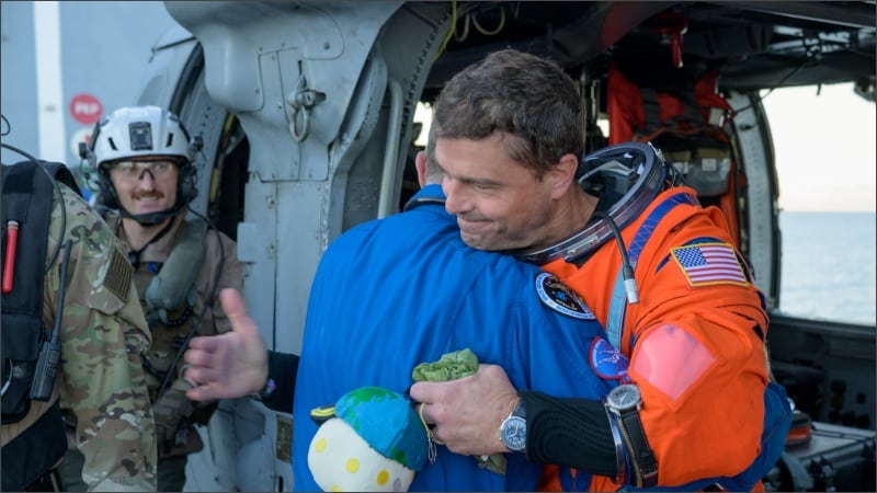 NASA astronaut Reid Wiseman in orange flight suit hugs flight surgeon Richard Scheuring on the flight deck of USS John P. Murtha after Artemis II splashdown in the Pacific Ocean