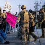 An armed federal ERO agent in tactical gear drags a limp protester by the arms outside the Broadview USCIS Processing Center in Illinois as demonstrators hold signs reading ICE GTFO of Illinois