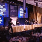 Tactical officers in body armor with rifles raised on the head table dais at the White House Correspondents' Dinner, with "Celebrating the First Amendment" visible on screens behind them