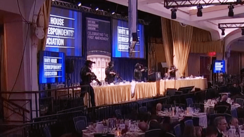 Tactical officers in body armor with rifles raised on the head table dais at the White House Correspondents' Dinner, with "Celebrating the First Amendment" visible on screens behind them