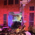 Nighttime view of a Katy, Texas brick home with a gaping hole in the front wall and debris scattered across the lawn after a Tesla crashed into it