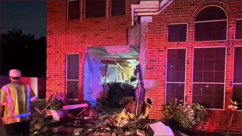 Nighttime view of a Katy, Texas brick home with a gaping hole in the front wall and debris scattered across the lawn after a Tesla crashed into it
