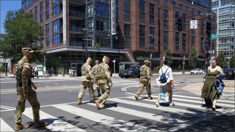 National Guard soldiers in camouflage cross a city street alongside civilian pedestrians near a Whole Foods in Washington, DC