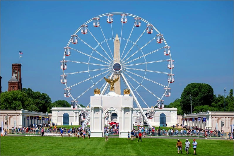 Ferris wheel encircling the Washington Monument behind a white triumphal arch at the Great American State Fair on the National Mall