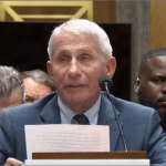 Anthony Fauci seated at a Senate hearing witness table, reading from a printed statement into a microphone.