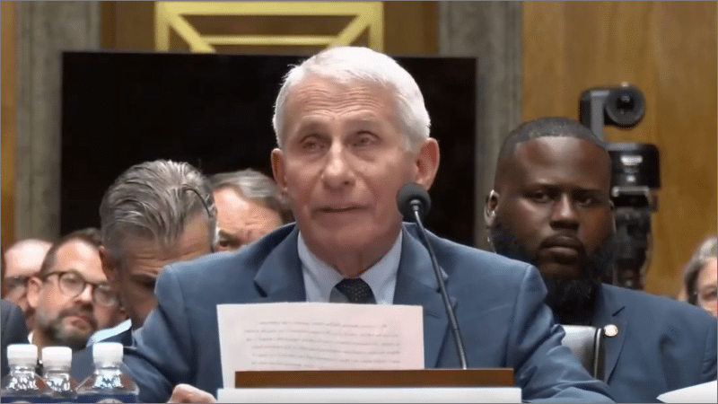 Anthony Fauci seated at a Senate hearing witness table, reading from a printed statement into a microphone.
