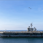 USS Abraham Lincoln aircraft carrier with rust-streaked hull at sea, full flight deck and two fighter jets overhead