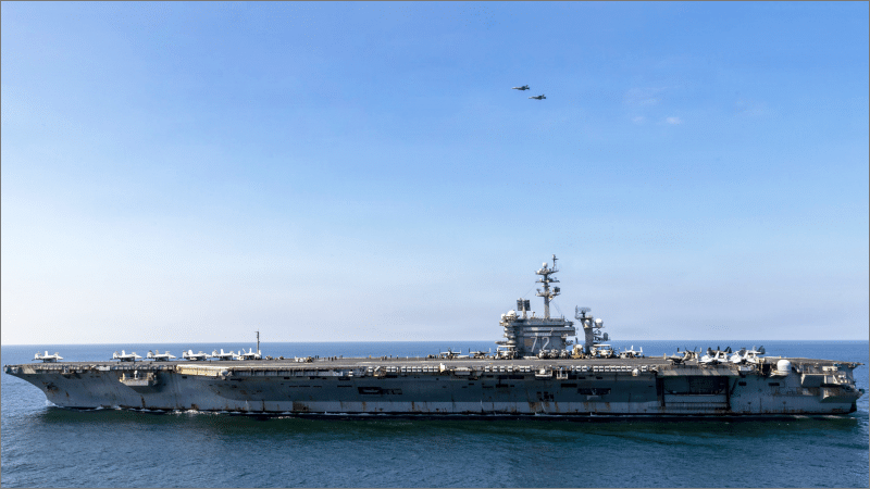 USS Abraham Lincoln aircraft carrier with rust-streaked hull at sea, full flight deck and two fighter jets overhead