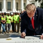 President Trump signs his name on the granite of the new White House helipad as construction workers in safety vests stand behind him with the White House in the background
