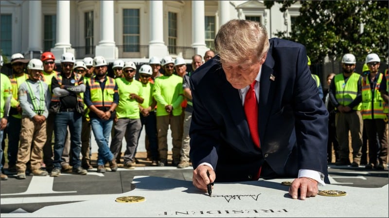 President Trump signs his name on the granite of the new White House helipad as construction workers in safety vests stand behind him with the White House in the background