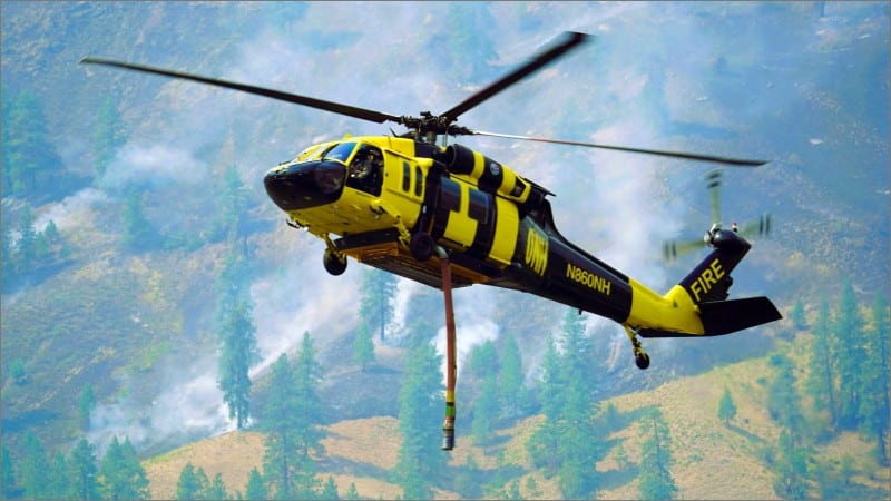 Yellow and black Sikorsky UH-60A Black Hawk firefighting helicopter carrying a water snorkel over smoke and forest at the Sinlahekin Fire in Washington