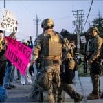 Masked ICE agents in tactical gear and camouflage confront protesters outside the Broadview ICE processing center, one demonstrator pulled to the ground beside a pink protest sign.