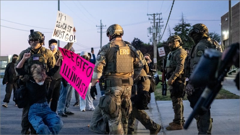 Masked ICE agents in tactical gear and camouflage confront protesters outside the Broadview ICE processing center, one demonstrator pulled to the ground beside a pink protest sign.