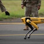 Yellow Boston Dynamics Spot robot dog walking on a runway between armed airmen in camouflage at RAF Leeming
