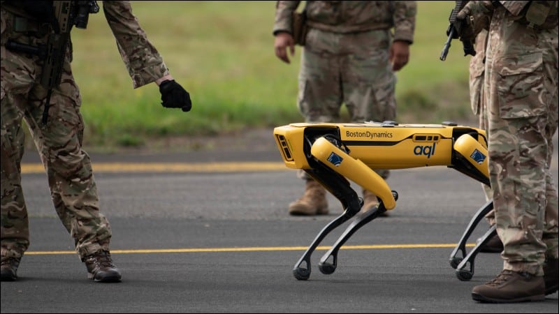 Yellow Boston Dynamics Spot robot dog walking on a runway between armed airmen in camouflage at RAF Leeming