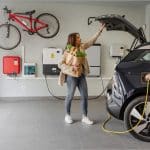 Woman holding groceries and boy plugging a charging cable into an electric car in a home garage