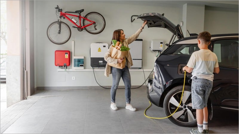 Woman holding groceries and boy plugging a charging cable into an electric car in a home garage