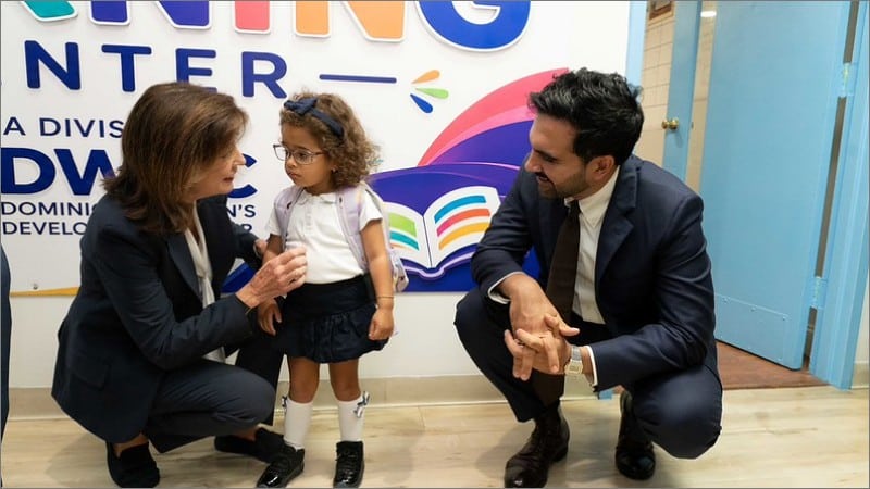 Gov. Kathy Hochul and Mayor Zohran Mamdani crouch beside a toddler in a school uniform at a Manhattan 2-K childcare provider on opening day.