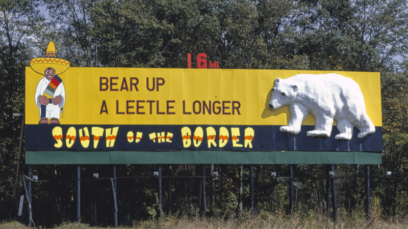 Yellow South of the Border billboard reading "Bear up a leetle longer, 16 mi" with a cartoon Mexican in a sombrero and a white polar bear