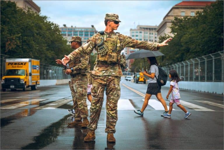 A National Guard soldier in camouflage directs traffic on a wet street near the National Mall as a woman with an umbrella and a young girl cross behind him.