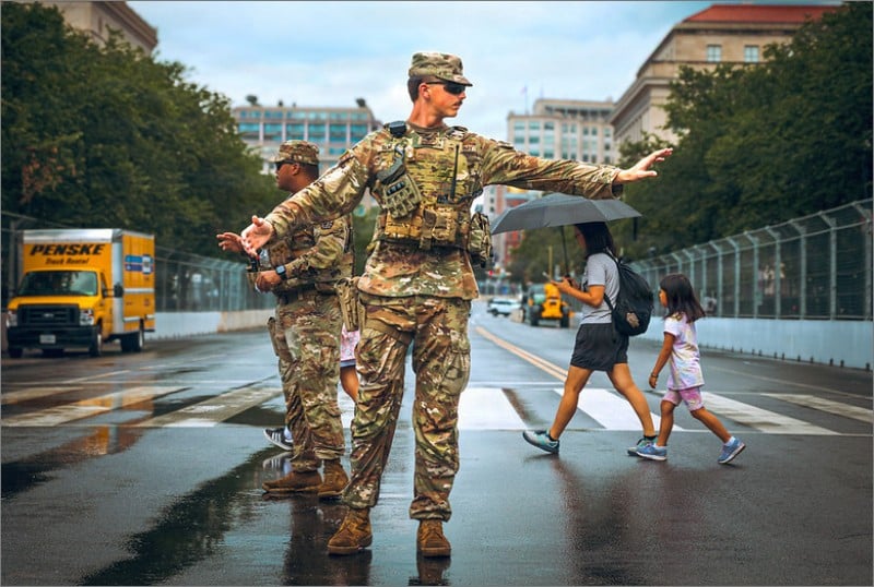 A National Guard soldier in camouflage directs traffic on a wet street near the National Mall as a woman with an umbrella and a young girl cross behind him.