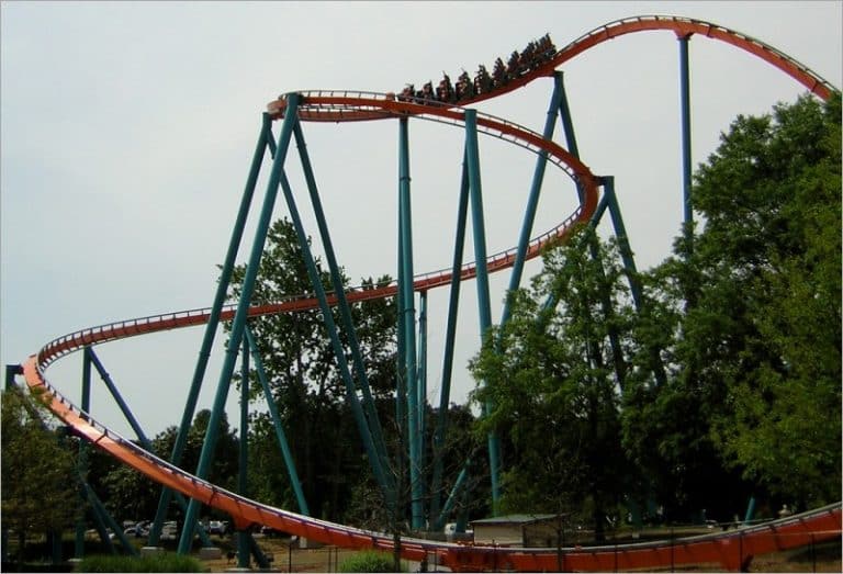 Goliath roller coaster at Six Flags Over Georgia, an orange steel track on teal supports curving above trees with a train of riders at the top
