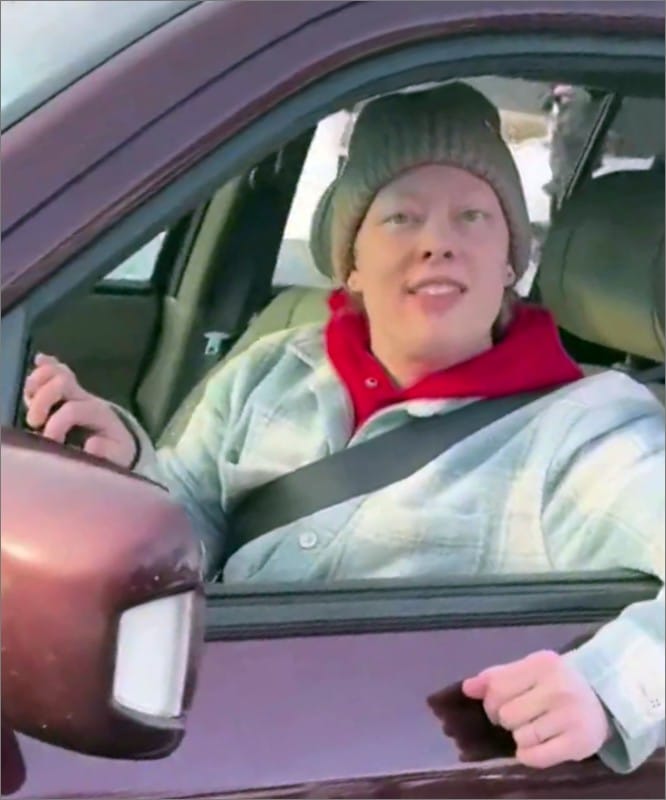 Renee Good, in a knit hat, red hoodie and plaid jacket, looks out the open driver's window of her maroon SUV at the ICE agent filming her in Minneapolis.