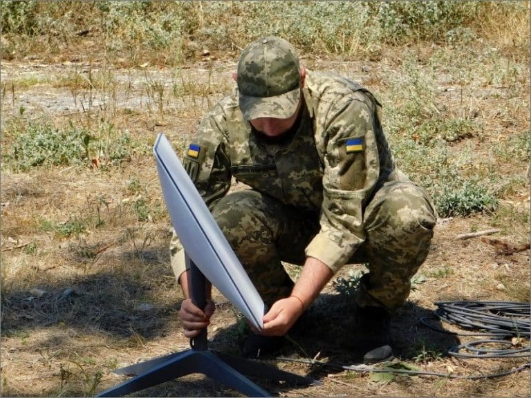 Ukrainian soldier in camouflage kneeling in a dry field to set up a white Starlink satellite dish on a metal stand