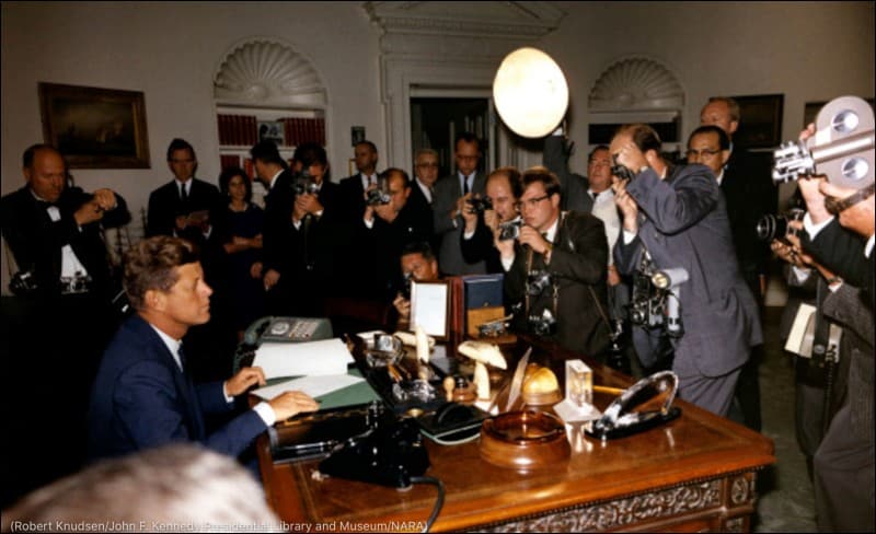 President John F. Kennedy seated at the Resolute desk in the Oval Office as photographers and reporters crowd around him in October 1962