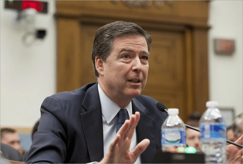 James Comey, in a dark suit and blue tie, raises his hand while testifying at a House Judiciary Committee hearing table with a microphone and water bottles.