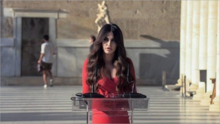 Kimberly Guilfoyle, U.S. ambassador to Greece, in a red dress at a clear podium with microphones beside white marble columns