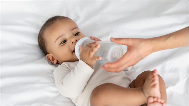 Infant in a cream bodysuit drinking from a bottle held by an adult hand while lying on a white bed