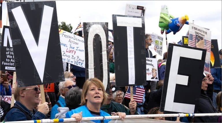 No Kings protesters at the Lincoln Memorial hold four large black signs spelling VOTE, with a crowd, handmade placards and an inflatable figure behind them.