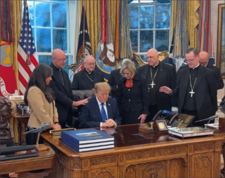 President Donald Trump sits with his head bowed at the Resolute Desk in the Oval Office as Catholic bishops stand behind him, some with hands on his shoulders, praying.